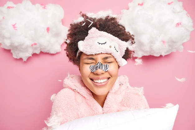 Positive young Afro American woman smiles toothily keeps eyes closed enjoys morning time after good sleep wears slumber suit blindfold on forehead holds pillow poses against flying feathers