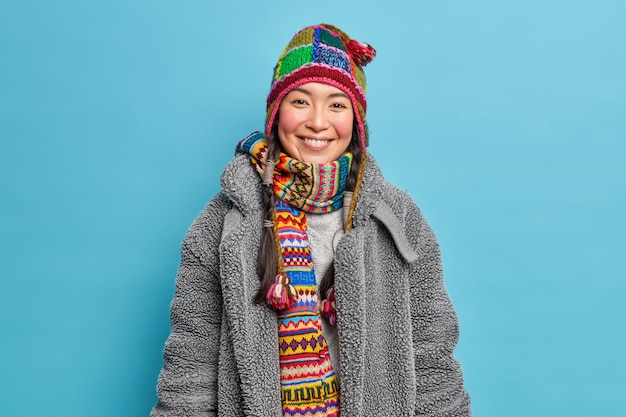 Free photo positive young asian woman with rouge cheeks smiles happily wears knitted hat and scarf around neck dressed in warm coat going to spend free time outdoors during winter day likes cold weather