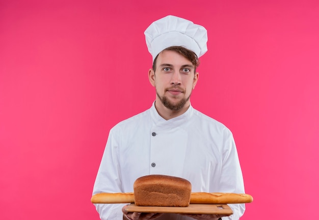 A positive young bearded chef man in white uniform holding a wooden kitchen board with several breads while looking on a pink wall