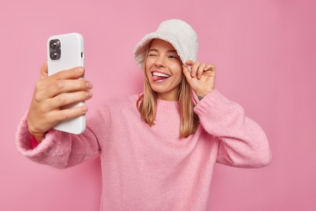 Positive young blonde woman winks eye and sticks out tongue makes selfie or video call via smartphone wears new panama loose jumper isolated over pink background foolishes around has upbeat mood