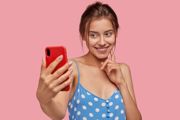 Positive young Caucasian woman takes picture of herself with modern cell phone, has tender smile on face, wears blue polka dor dress, models against pink wall. Pretty lady poses for selfie