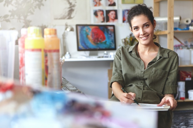 Free photo positive young creative woman dressed casually, sitting at her workshop, making sketches with pencil, being involved in creative process, enjoying her work. people, lifestyle and art concept