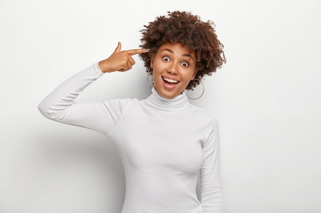 Positive young curly woman holds index finger on temple, tilts head, has joyous face expression, wears round earrings and poloneck