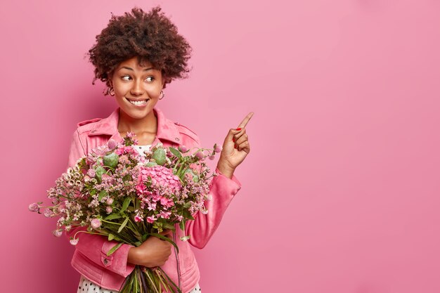 Positive young curly woman poses with nice bouquet of flowers points at blank space shows advertising content wears jacket isolated over pink wall