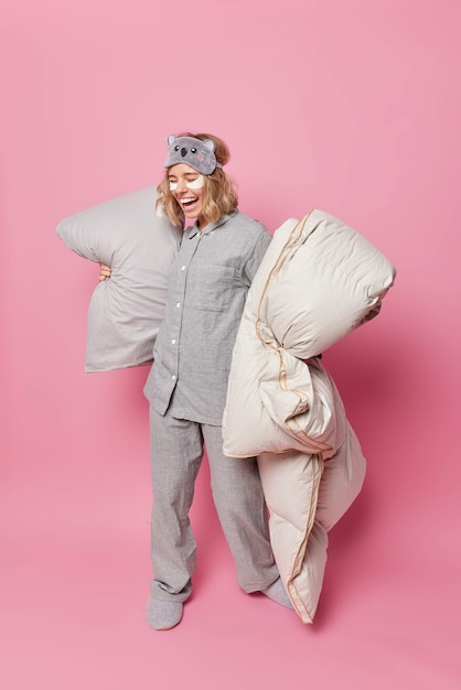 Positive young European woman dressed in grey nightwear holds pillow and blanket laughs joyfully applies beauty patches under eyes goes to bed isolated over pink background. Full length shot