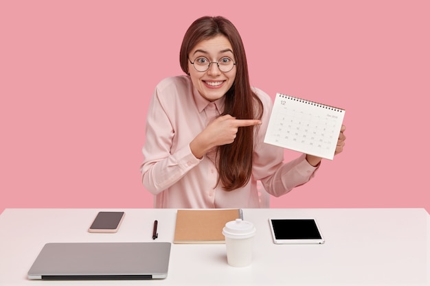 Free Photo positive young freelance worker points at calendar, demonstrates date of finishing project work, dressed in fashionable clothes, poses at desktop