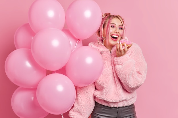 Positive young teenage girl with bright pink makeup bites delicious sweet doughnut