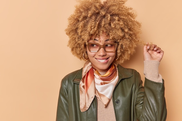 Positive young woman curls hair bites lips and looks away being in good dreams about something wears spectacles fashionable leather jacket focused away poses against beige background copy space