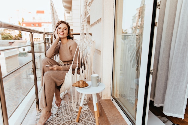 Free photo positive young woman in long dress sitting at balcony with coffee and croissant. photo of barefooted curly girl enjoying breakfast at terrace.