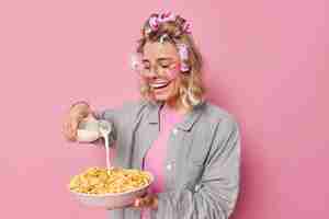 Free photo positive young woman makes hairstyle with rollers going to have breakfast pours milk in cornflakes dressed in nightwear undergoes beauty procedures isolated over pink background morning time