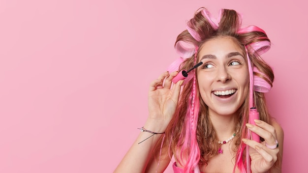 Free Photo positive young woman puts on mascara applies hair rollers for making curly hairstyle smiles broadly wants to have fabulous look concentrated away poses against pink background with copy space