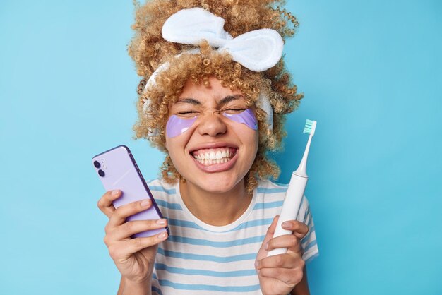 Positive young woman shows white teeth after cleaning takes care of skin applies bauty patches holds modern electric toothbrush and smartphone wears headband striped t shirt isolated on blue wall