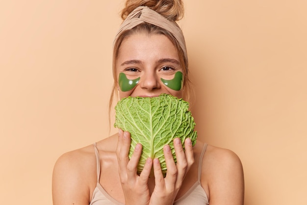 Free photo positive young woman uses natural products for skin care holds cabbage over mouth wears green hydrogel patches under eyes wears headband and t shirt isolated on beige background. beauty time