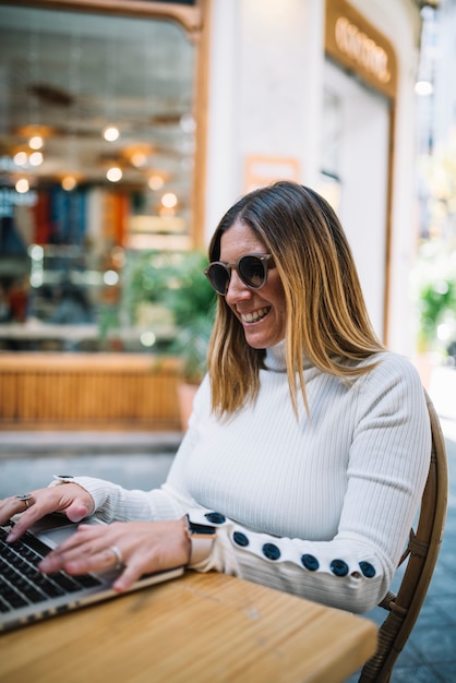 Positive young woman using laptop at table in street cafe