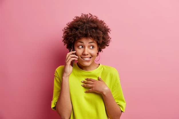 Free photo positive young woman with curly hair talks via mobile phone bites lips looks aside feels glad dressed in casual t shirt isolated over pink wall waits until friend will answer