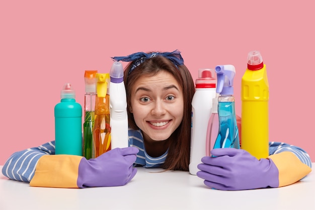 Positive young woman with delighted expression, toothy smile, embraces chemical detergents, looks gladfully 