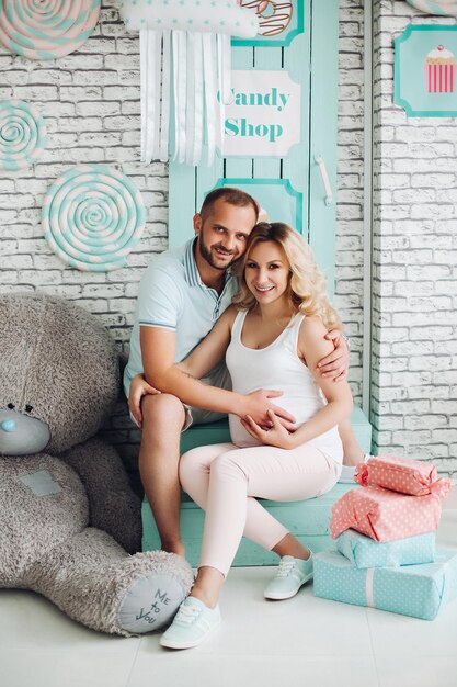 Positivity couple of handsome man and pretty blonde pragnant woman sitting on stairs at studio embracing stomach and gesturing heart by hands Young stylish family at sweet mint interior among toys