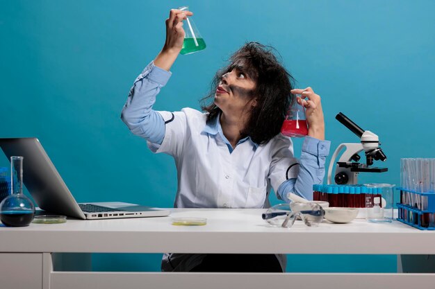 Potrait of crazy silly chemist analyzing glass jars filled with liquid experiment serums while sitting at desk. Foolish goofy looking maniac scientist having beakers filled with chemical compounds.