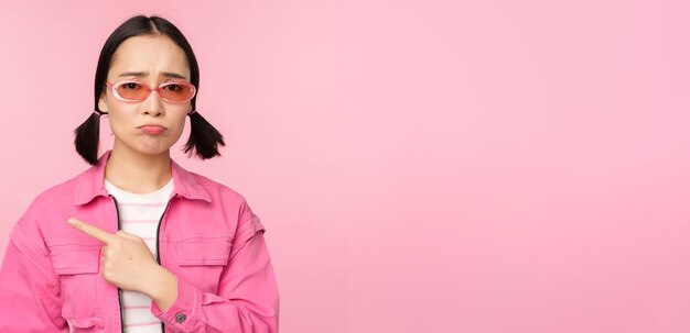 Potrait of cute korean girl in sunglasses pointing left and looking disappointed sulking upset standing over pink background