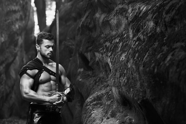 Free Photo power in peace. monochrome shot of a calm thoughtful warrior standing with a sword near the rocks at the woods. young strong man with a muscular torso posing copyspace