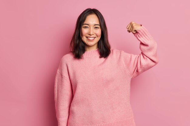 Powerful cheerful brunette woman raises arm and shows muscle demonstrates her strength looks confident at camera smiles gently wears casual long sleeved jumper 