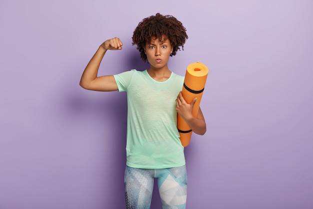 Powerful strong woman raises arm, shows biceps, holds fitness mat for gym training, dressed in active wear