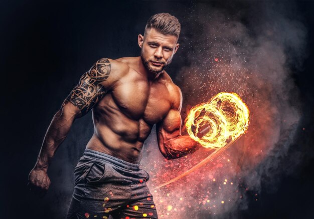 Powerful stylish bodybuilder with tattoo on his arm, doing the exercises on a biceps with dumbbells, look at the camera with a confident look. Fire art concept.