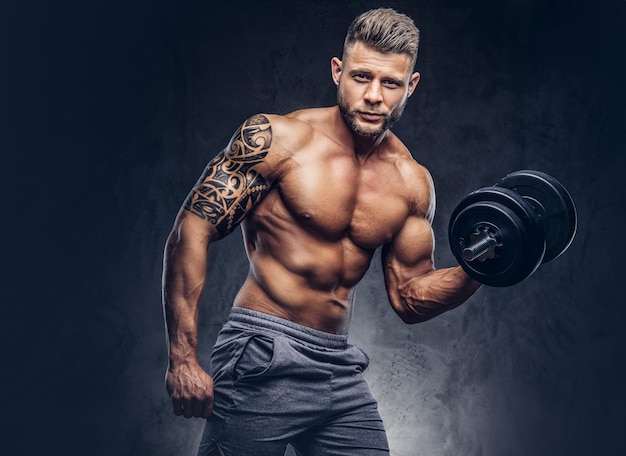 Free photo powerful stylish bodybuilder with tattoo on his arm, doing the exercises on a biceps with dumbbells, look at the camera with a confident look. isolated on a dark background.