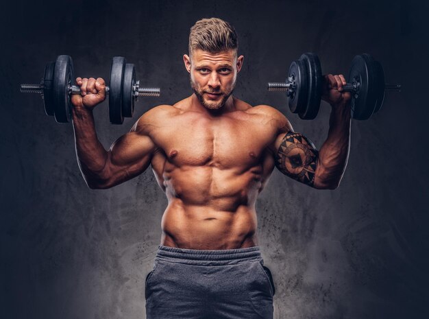 Powerful stylish bodybuilder with tattoo on his arm, doing the exercises with dumbbells. Isolated on a dark background.