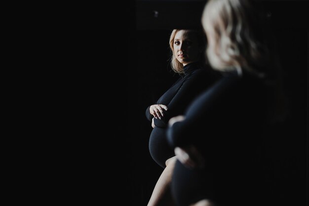 Pregnant blonde woman in a black bodysuit stands near a mirror and looks at her reflection