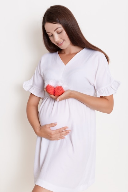 Free photo pregnant happy woman holding red knitted baby shoes in her hands, touching her belly with charming smile