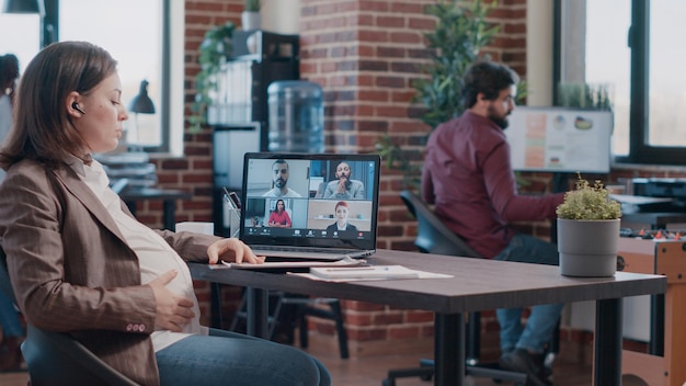 Free photo pregnant woman talking to colleagues on video call, using laptop at office. employee expecting child and attending business meeting with workmates on video conference to talk about project