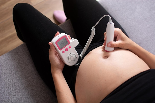 Free Photo pregnant woman with a fetal doppler listening to the baby's heart sitting on the sofa at home