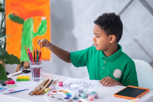 Preparation, drawing. Profile of dark-skinned involved boy in green tshirt taking out pencil from glass sitting at table preparing to draw in daylight