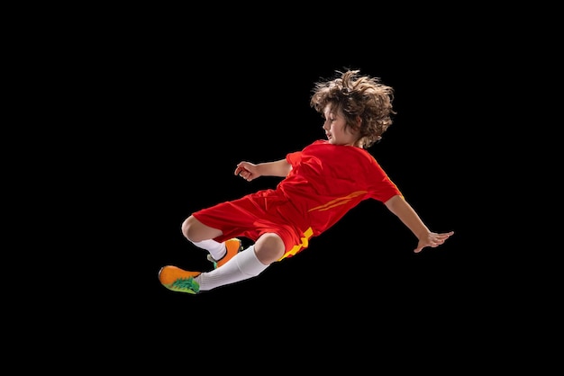 Preschool boy football soccer player in red sports uniform practicing with football ball isolated on dark studio background Concept of sport game hobby