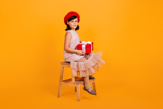 Preteen girl in dress holding birthday gift.  kid with present sitting on chair on yellow wall.