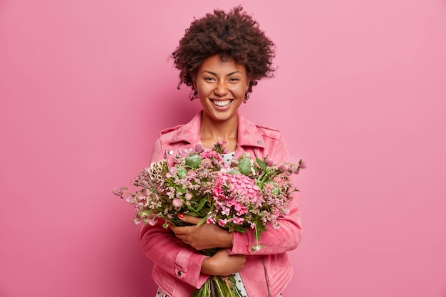 pretty African American woman expresses sincere emotions, embraces bouquet of flowers, has spring mood