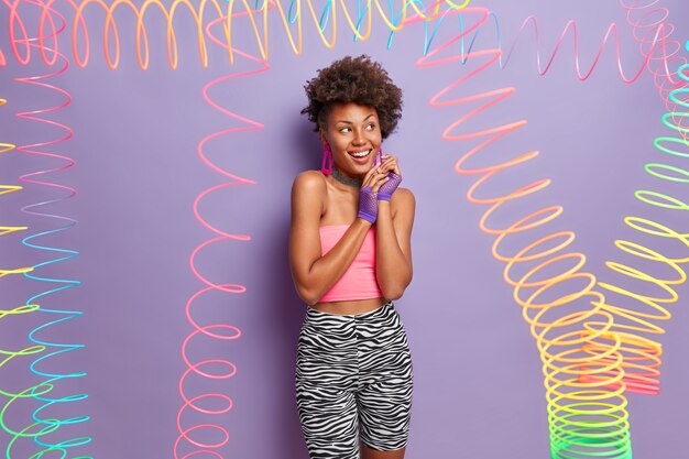 Pretty African American woman keeps hands together, looks aside, smiles broadly, dressed in casual active wear