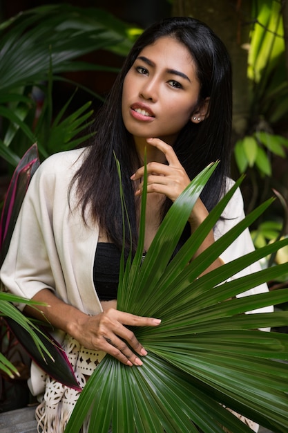 Pretty asian woman posing in tropical garden, holding big palm leaf.