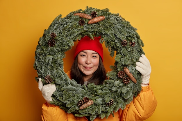 Free photo pretty asian woman wears warm headgear and gloves, looks through handmade traditional christmas wreath, being in good festive mood, anticipates for winter holidays, isolated on yellow studio wall