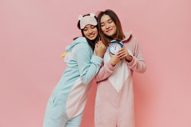 Pretty Asian women in pajamas pose on pink background Cute young girl in kigurumi smiles and holds blue alarm clock