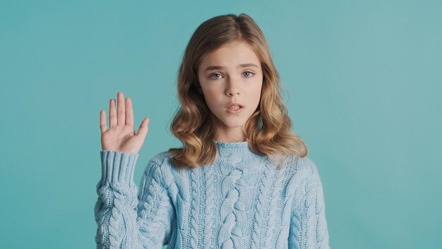 Pretty blond wavy haired teenage girl waving goodbye to friend looking sad isolated on blue background Bye bye expression