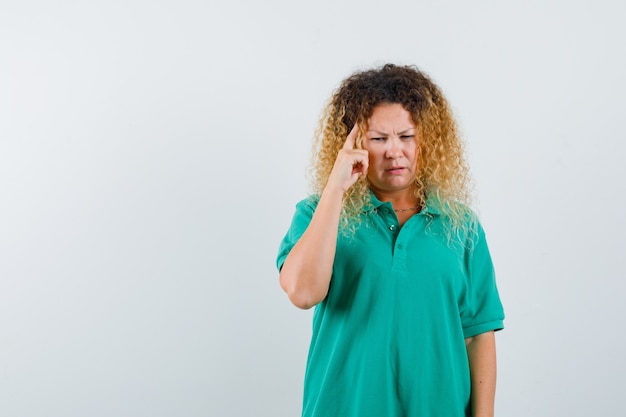 Pretty blond woman keeping finger on temples in green polo t-shirt and looking thoughtful. front view.