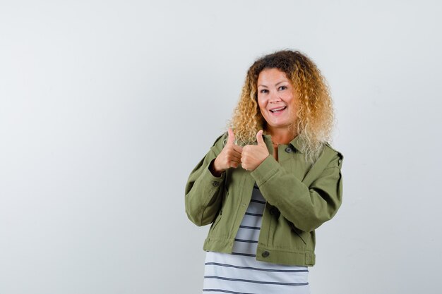 Pretty blond woman showing double thumbs up in green jacket and looking merry. front view.