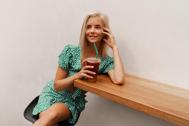 Pretty blond woman sitting in modern cafe and drinking sweet lemonade Wearing stylish dress