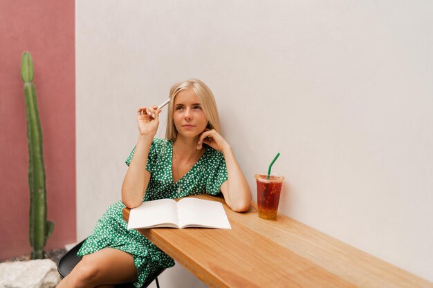 Pretty blond woman writing in notebook Wearing summer dress Sitting in modern cafe with white walls