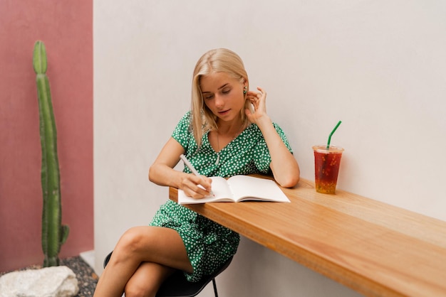 Pretty blond woman writing in notebook Wearing summer dress Sitting in modern cafe with white walls