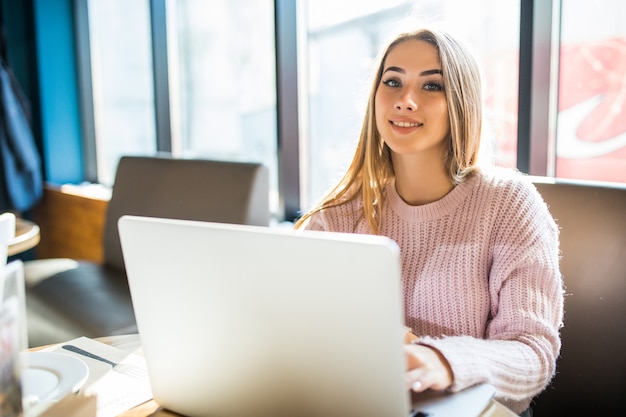 Pretty blonde girl in fashion white sweater in working on her laptop computer in cafe in daily time