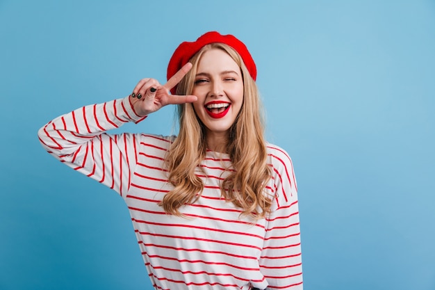 Free Photo pretty blonde girl in striped shirt showing peace sign. front view of laughing french lady posing on blue wall.