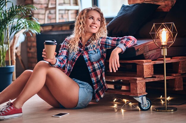 Pretty blonde hipster female drinks morning coffee in a restroom with loft interior.